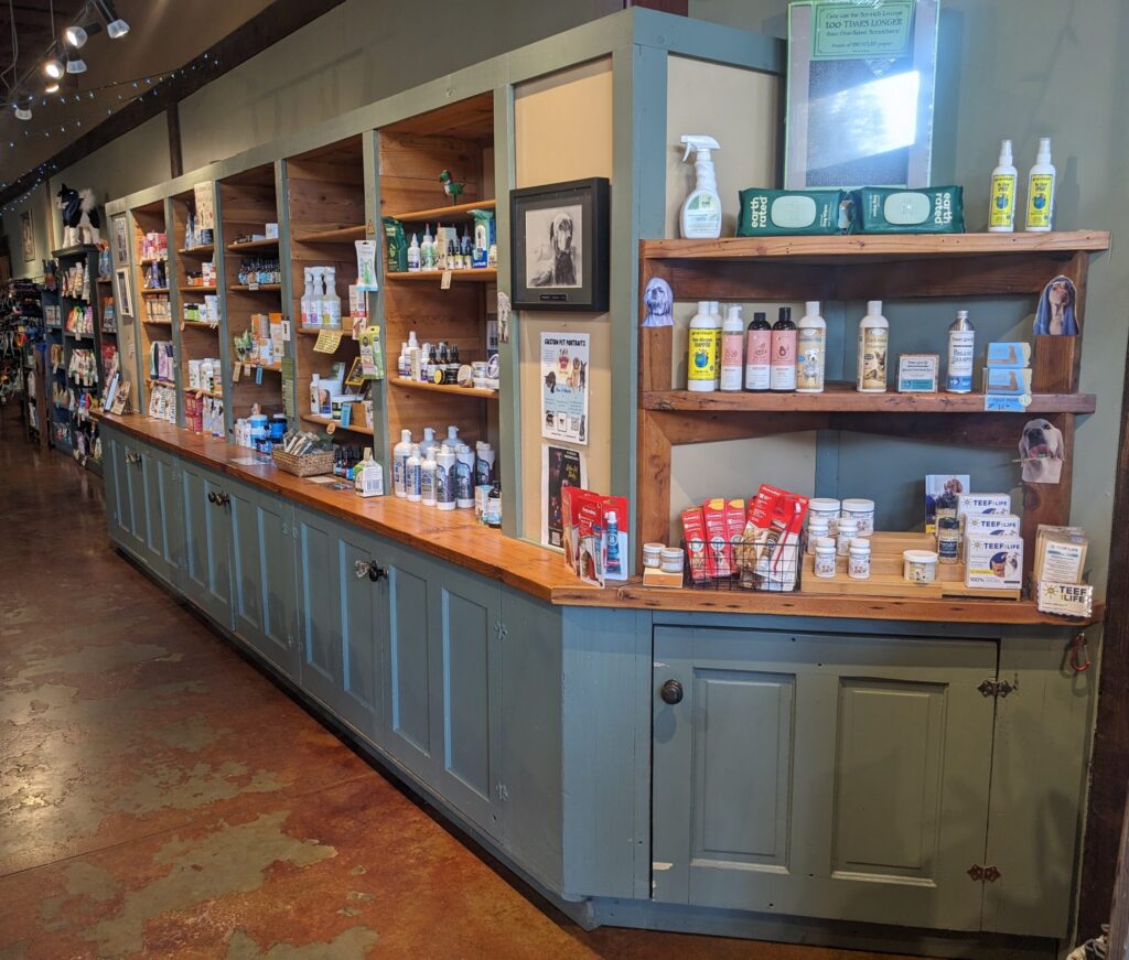 Light green shelving attached to the wall is filled with pet supplies in the new store location. A dark brown wooden border is visible in the background of the photo.