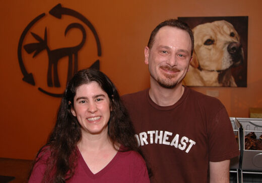 Christine and Mike stand together smiling at the camera in front of an orange wall with the Green Dog Logo and a portrait of a yellow lab on it.